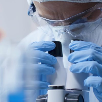 Closeup of scientist doctor looking into medical microscope analyzing blood sample during scientific experiment in biochemistry laboratory. Researcher man developing vaccine against covid19